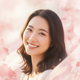 A smiling young adult woman of East Asian descent, looking happy and trustworthy against a soft pink and white background.