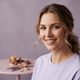 Headshot of a smiling woman in her 30s with light brown hair and a soft lavender top, against a blurred dusty lilac backgroun